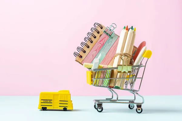 Buying school supplies, pencils, tape, notebook and crayons. Stationery for kids in a shopping cart with yellow school bus on pink turquoise background, copy space. Back to school concept.