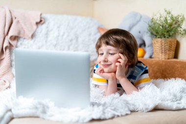 Okula dönelim. Uzaktan öğrenme çevrimiçi eğitim. Beyaz tebessümlü anaokulu çocuğu evde dizüstü bilgisayarla ders çalışıyor ve ödev yapıyor. Kanepe ve battaniyeye uzanmış bilgisayarla