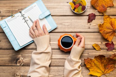 Kadın eli süveter, balkabağı stili kahve ve sonbahar temalı süslemeler, düşmüş yapraklar ve ahşap arka planda bir defter. Düz yat, boşluğu kopyala