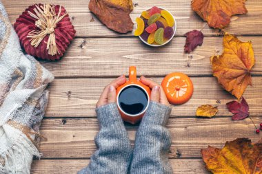 Gri kazaklı kadın elleri, balkabağı stili kahve ve sonbahar temalı süslemeler, düşmüş yapraklar ve ahşap arka planda şekerler. Düz yat, boşluğu kopyala.