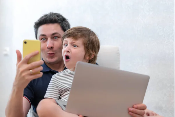 令人惊讶的年轻人坐在电脑上和学龄前的儿子一起使用笔记本电脑放松一下 爸爸和小男孩一起享受家里的智能手机 坐在椅子上忙着玩具娃娃 网上教育 — 图库照片