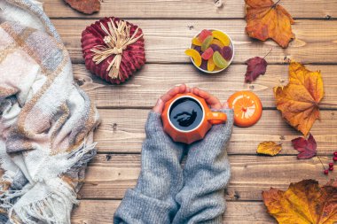 Gri kazaklı kadın elleri, balkabağı stili kahve ve sonbahar temalı süslemeler, düşmüş yapraklar ve ahşap arka planda şekerler. Düz yat, boşluğu kopyala.