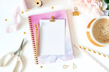 Masaüstü modeli, el yapımı DIY araçları. Beyaz çalışma masasının arka planında bir fincan kahve. Düz makas, bant, planlayıcı, altın kağıt ataçları, boş tebrik kartı, not defteri ve kalem..
