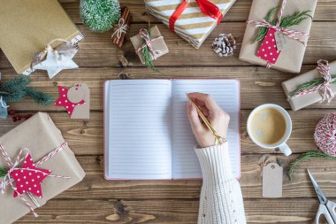 Masaüstü planlayıcısı. Bir deftere bir kadın eli yazıp Noel hediyesini paketliyor. Dekor unsurlarıyla ahşap arka planda sunumlar, üst görünüm. Yeni yıl paketleme kavramı, üst görünüm