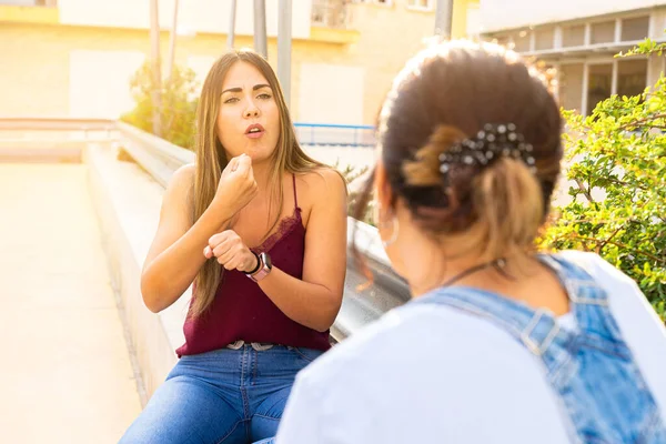 Two girls speaking in sign language in a public space. Deaf friends or couple communicating, having fun, pleasant conversation, sitting together