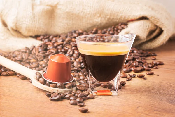 Cup of coffee with coffee capsule on wooden spoon, roasted coffee beans on wooden background,front view.