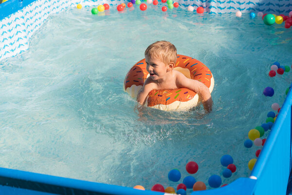 Happy baby boy playing with colorful inflatable ring in outdoor small swimming pool on hot summer day. Kids learn to swim. Child water toys.