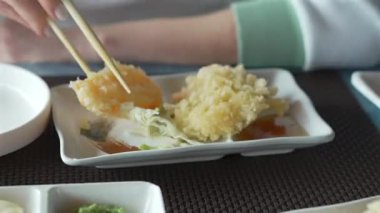 Elinde çubuk olan bir kadın ince dilimlenmiş Japon sebze tempurası ve zencefilli ponzu soslu sağlıklı vejetaryen yemeği alır. Restoran masasında servis edilen lezzetli Asya yemeği.