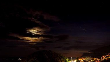 Deniz üzerinde bulutların arasından batışı. Küçük bir şehrin gece ay ayarına. Vurulmuş trafik ve gets geç gidiyor ışıkları ile hızlandırılmış. Katic ve Adriyatik Denizi, Karadağ, Petrovac, Lucice Sveta şeridinde Adaları. 4k, 3840 x 2160, Timelapse