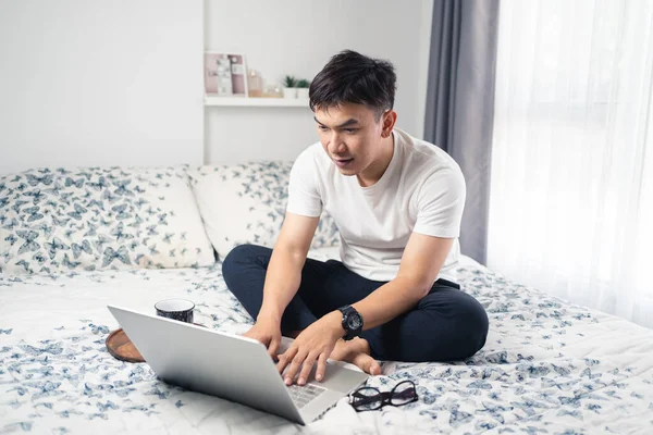 Young asian working at home. A man sitting on bed and working with laptop computer on bed. Work from home concept, Asia