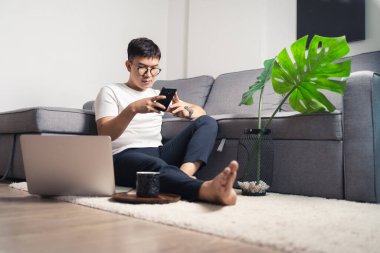 Genç Asyalı adam evde çalışıyor ve bilgisayarla çalıştıktan sonra akıllı telefonla sohbet ediyor. Ev kavramından çalış. Bangkok Tayland