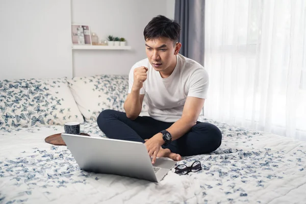 Young Asian man looking at laptop computer excited by good news online. Successful with a big project while working at home. Work from home concept. Bangkok Thailand