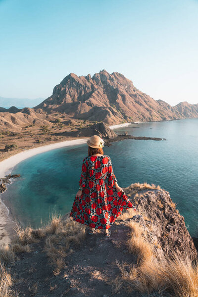 Young woman traveller wearing red dress standing on top of Padar island in a morning sunrise, Flores island in Komodo national park, Indonesia, Asia