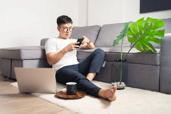 Young Asian man chatting with smartphone after working by using laptop computer. A man relaxing and sitting on floor in private room. Work from home concept. Bangkok Thailand
