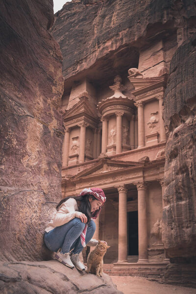 Молодой азиатский путешественник с котом сидит на скале перед казначейством или или Аль-Хазне в руинах Петра и древнем городе Набатея королевства, Иордании, арабских, Азии
