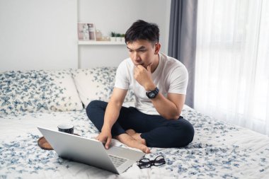 Genç Asyalı adam dizüstü bilgisayara bakıyor ve evde çalışırken bir şeyler düşünüyor. Yatakta oturan gözlüklü bir adam. Ev kavramından çalış. Bangkok Tayland