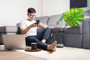 Genç Asyalı adam bilgisayarla çalıştıktan sonra akıllı telefonla sohbet ediyor. Rahatlayan ve özel odada yerde oturan bir adam. Ev kavramından çalış. Bangkok Tayland