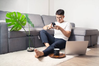 Genç Asyalı adam bilgisayarla çalıştıktan sonra akıllı telefonla sohbet ediyor. Rahatlayan ve özel odada yerde oturan bir adam. Ev kavramından çalış. Bangkok Tayland