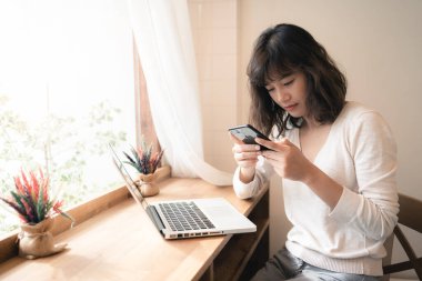 Genç Asyalı kadın dizüstü bilgisayarla çalışırken akıllı telefonu sohbet etmek için kullanıyor. Evde çalışırken başka insanlarla iletişim kuran bir kadın. Sosyal uzaklık kavramı. Bangkok, Tayland