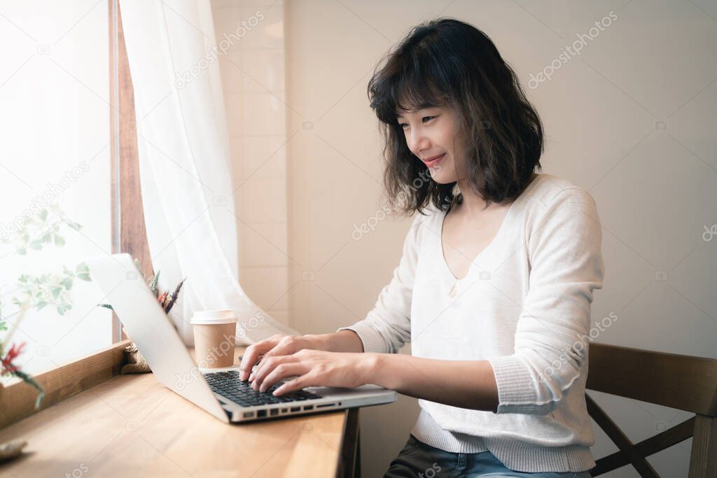 Young Asian woman working and video conferencing with laptop computer. A happy woman with smiley face working from home. A cup of coffee on wooden table. Social distancing concept. Bangkok, Thailand
