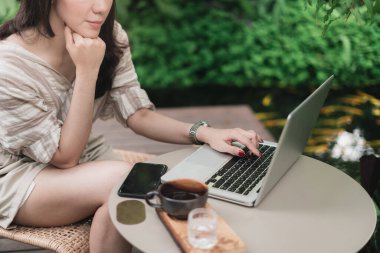 Bilgisayarı kullanan, akıllı telefon ve bir fincan kahveyle bahçede internetten bilgi arayan kadınların ellerini kapat. Teknoloji sektörü ve sosyal uzaklık kavramı. Bangkok, Tayland