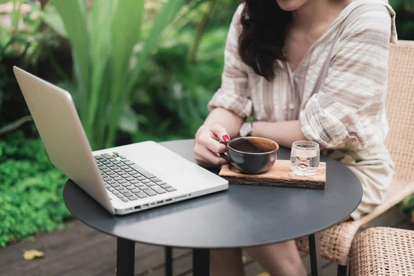 Close up hands of woman using laptop computer working and searching online information in garden with cup of coffee on a table. Technology business and social distancing concept. Bangkok, Thailand