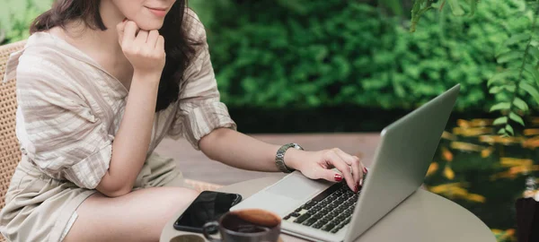 Close up hands of woman using laptop computer working and searching online information in garden with smartphone and cup of coffee on table. Technology business and social distancing concept. Banner portion. Thailand