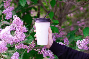 Doğanın, çiçeklerin ve leylağın arka planında plastik bir bardak kahveyle kadın eli.