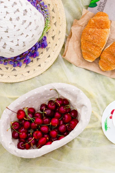 Yaz - çayırda piknik. Tatlı kiraz, kahve, içkili kupa, çiçekler, lupinler, kruvasanlar, çörekler, tatlı, şapka, sepet.