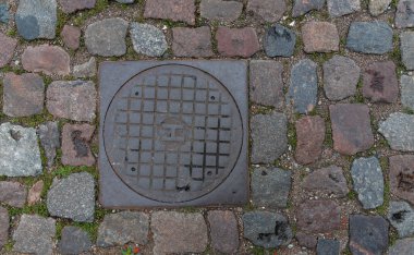 Metal rögar kapağı. Yol granit kaldırım taşlarıyla döşenmiş. Kaldırımda eski inşaatlar.