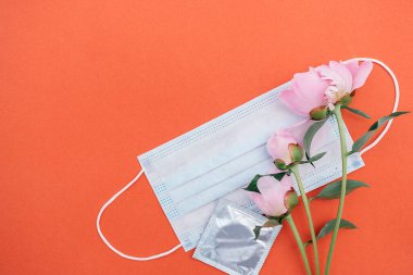 Koruyucu tıbbi maske, pembe şakayık tomurcukları ve kırmızı arka planda bir prezervatif. Yeni normal gerçeklikte aşk ilişkisi kavramı. Uzayı kopyala, düz yat