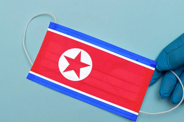 The flag of North Korea on a medical mask in the hands of a doctor on a blue background.Coronavirus Covid-19 pandemic concept