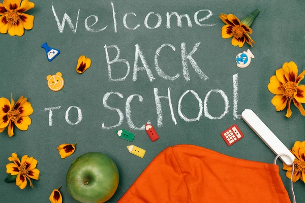 Blackboard with the inscription WELCOME BACK TO SCHOOL,chalk,apple, flowers and protective face mask.Back to school concept in a new coronavirus reality.New normal concept.Flat lay,top view