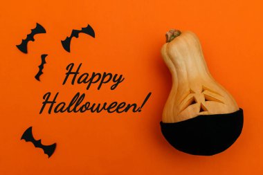 Siyah koruyucu tıbbi maskeli Cadılar Bayramı kabağı ve turuncu arka planda siyah kağıttan yarasalar. Yazı: Mutlu Cadılar Bayramı! Hem de turuncu bir arka planda. Cadılar Bayramı ve covid-19 kavramı..