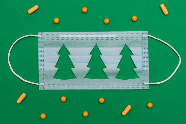 Medical protective mask with a cut-out silhouettes of a Christmas tree on a green background.Yellow pills are scattered on the background.Christmas, Happy New Year,coronavirus and medical concept.