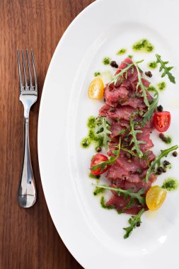 Et carpaccio, beyaz bir yerde çiğ et, domates ve otlar, çatallı roka, yukarıdan. Beyaz tabakta roka ve parmesan peynirli et carpaccio. en üstteki görünüm.