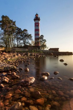Leningrad bölgesindeki Karavaldai yarımadasının ucundaki Shepelevsky deniz feneri bir yaz akşamı taş plajın ortasında gün batımında.