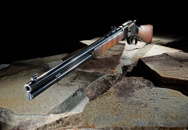 Lever action rifle like the ones used in the old west with a wood stock and blue steel on stones outdoors with a dark background.