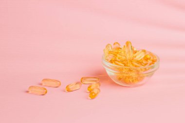 Balık yağı takviyesi kapsülleri küçük bir cam kaseden pembe bir yüzeye dökülüyor, sağlık ve refahı arttırıyor.