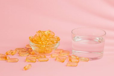 Cam kaseden dökülen balık yağı kapsülleri ve pembe arka planda duran bir bardak su, sağlıklı bir yaşam tarzını temsil ediyor.