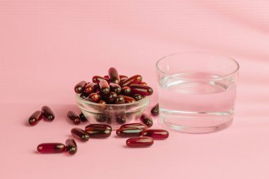 Kahverengi iyot takviyesi hapları cam kaseden dökülüyor ve pembe arka planda bir bardak su, sağlık ve sağlığı destekliyor.