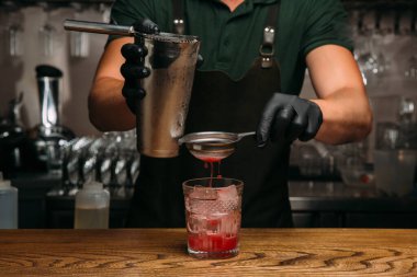 Barmen siyah eldiven giyiyordu. Titreştiriciden süzülen cama kırmızı kokteyl döküyordu. Tahta tezgahın üzerinde buzla birlikte.