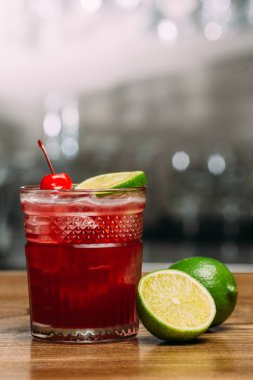 Kiraz ve kiraz dilimiyle süslenmiş canlı kırmızı kokteyl, arka planı bulanık, ahşap bir tezgahta oturuyor.