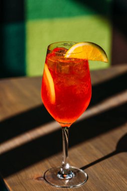 Parlak kırmızı aperol sprey kokteyli, portakal dilimi ve buz küpleri, parlak güneş ışığında ahşap masanın üzerinde durmak, ferahlatıcı yaz titreşimleri yaratıyor.
