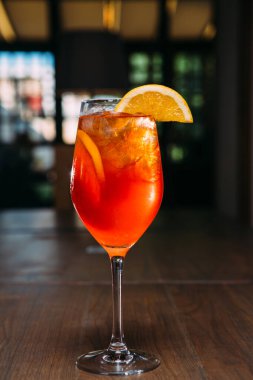 Taze portakal dilimi ve buz küpleriyle süslenmiş ahşap bir masada duran canlı spritz kokteyli canlandırıcı ve davetkar bir sahne yaratıyor.