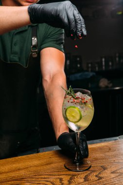 Barmen siyah eldiven giyiyor. Kokteyl bardağına buz, limon, biberiye ve karışık bir içecek dolu pembe biber ekliyor.