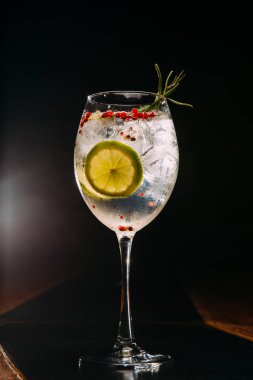 Limon tekeri, biberiye filizi ve kırmızı biberiyeler ile süslenmiş cin tonik kokteyl, koyu bir arka planda zarif bir bardakta servis edilir.