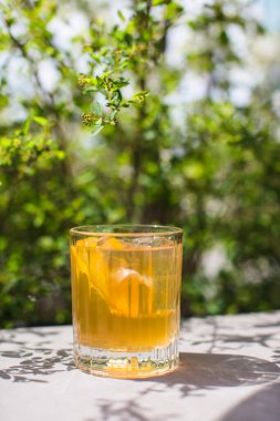 Glass of amber drink with citrus slices and ice on a table, with green foliage blurring in the background
