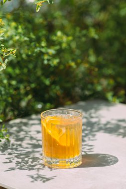 Refreshing orange drink with ice cubes and orange slice on a gray table outdoor in bright sunny day. Green foliage in background