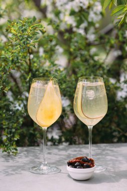 Two cocktails with pear slices and ice on a table, creating a summer vibe, with a bowl of olives and dried tomatoes